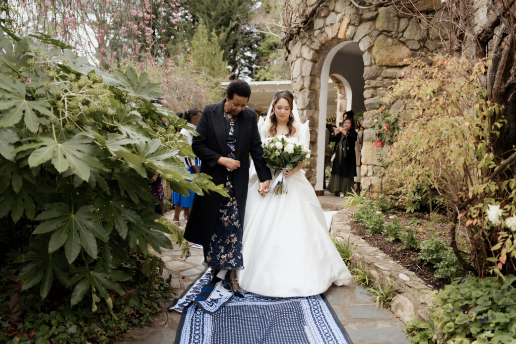 Bride Karen being joyfully escorted to the ceremony by a group of African women, singing and clapping as they walk through the gardens at The Manor Basket Range.