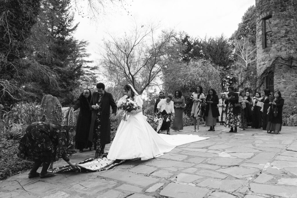 Bride Karen being joyfully escorted to the ceremony by a group of African women, singing and clapping as they walk through the gardens at The Manor Basket Range.